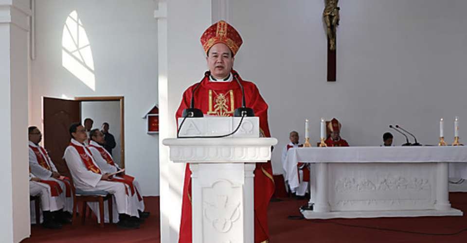 A bishop delivers a sermon during a religious ceremony in a church, with clergy members seated in the background.