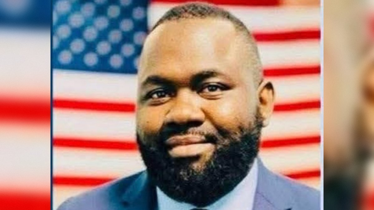 Portrait of a smiling man with a beard, dressed in a suit, in front of an American flag background.