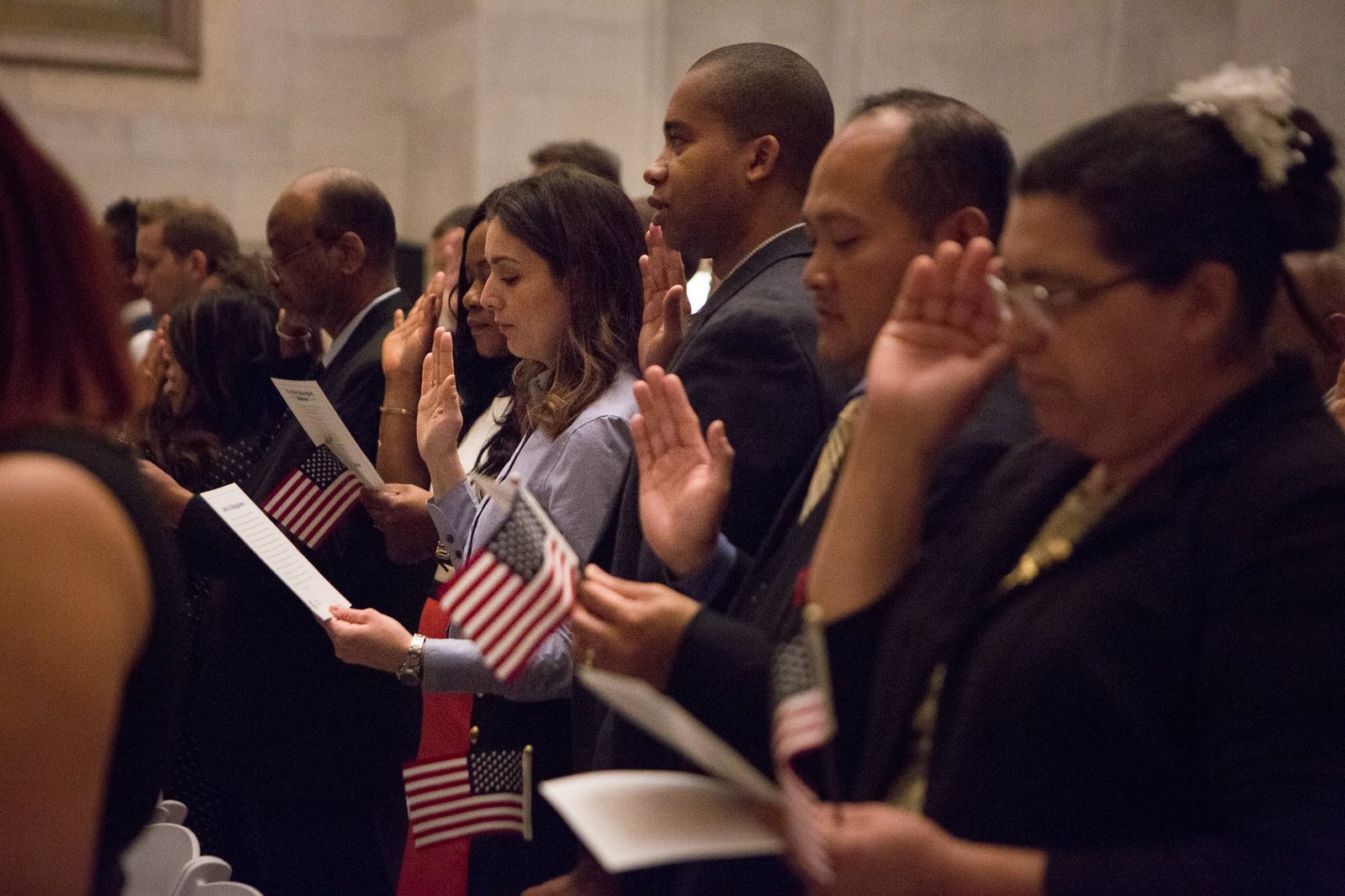 Diverse group of new citizens taking the Oath of Allegiance while holding small American flags during a naturalization ceremony.