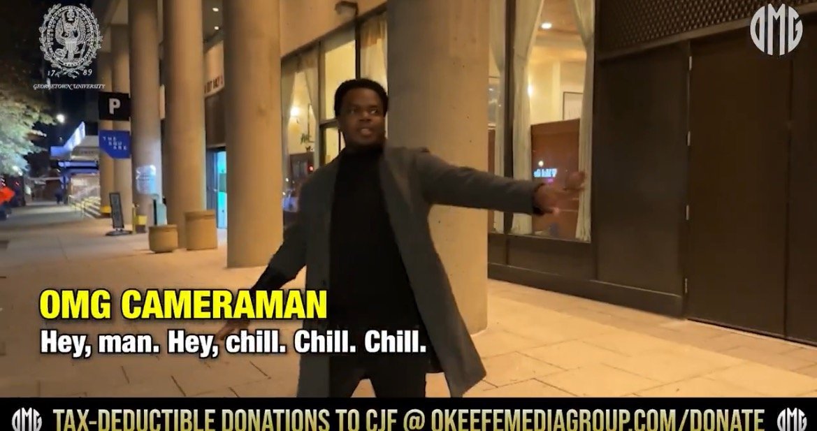 A man in a coat gestures while speaking on a city street at night, with storefronts and columns in the background.