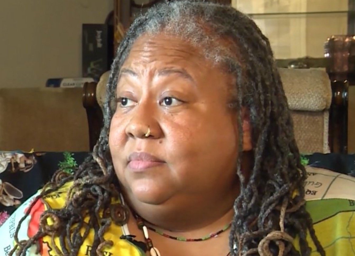 Close-up of a woman with dreadlocks and a colorful shirt, sitting on a couch, looking thoughtfully to the side in a cozy indoor setting.