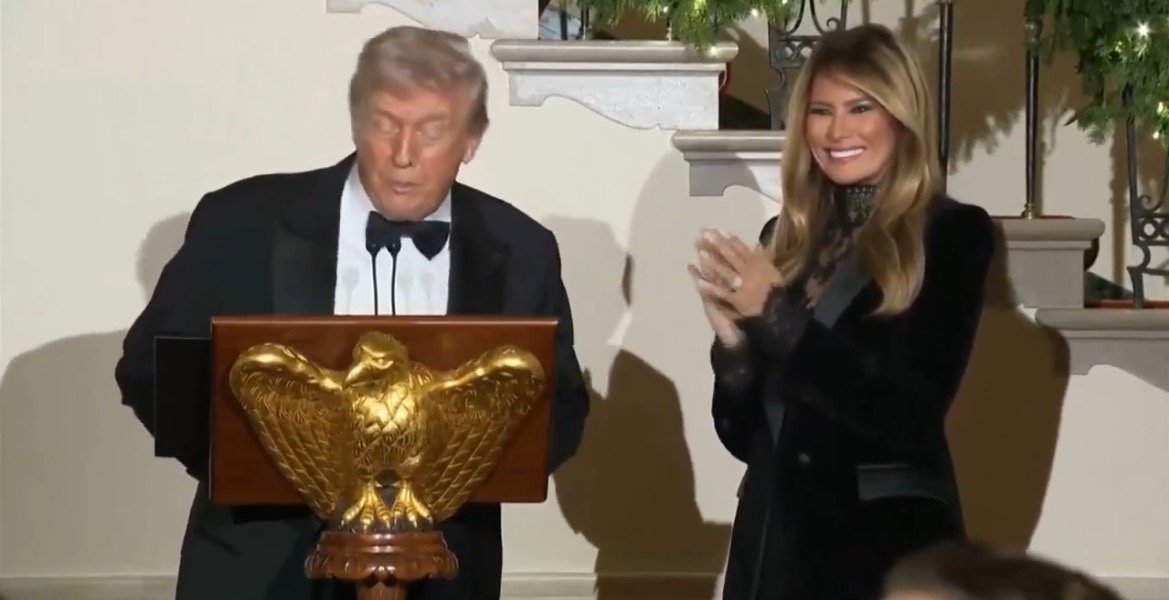 Donald Trump speaks at an event while Melania Trump applauds, with an eagle emblem visible on the podium.
