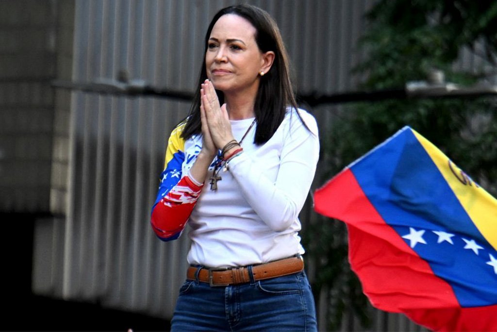 Venezuelan activist stands with hands clasped in prayer, wearing a shirt featuring the national flag colors, as a Venezuelan flag waves in the background.