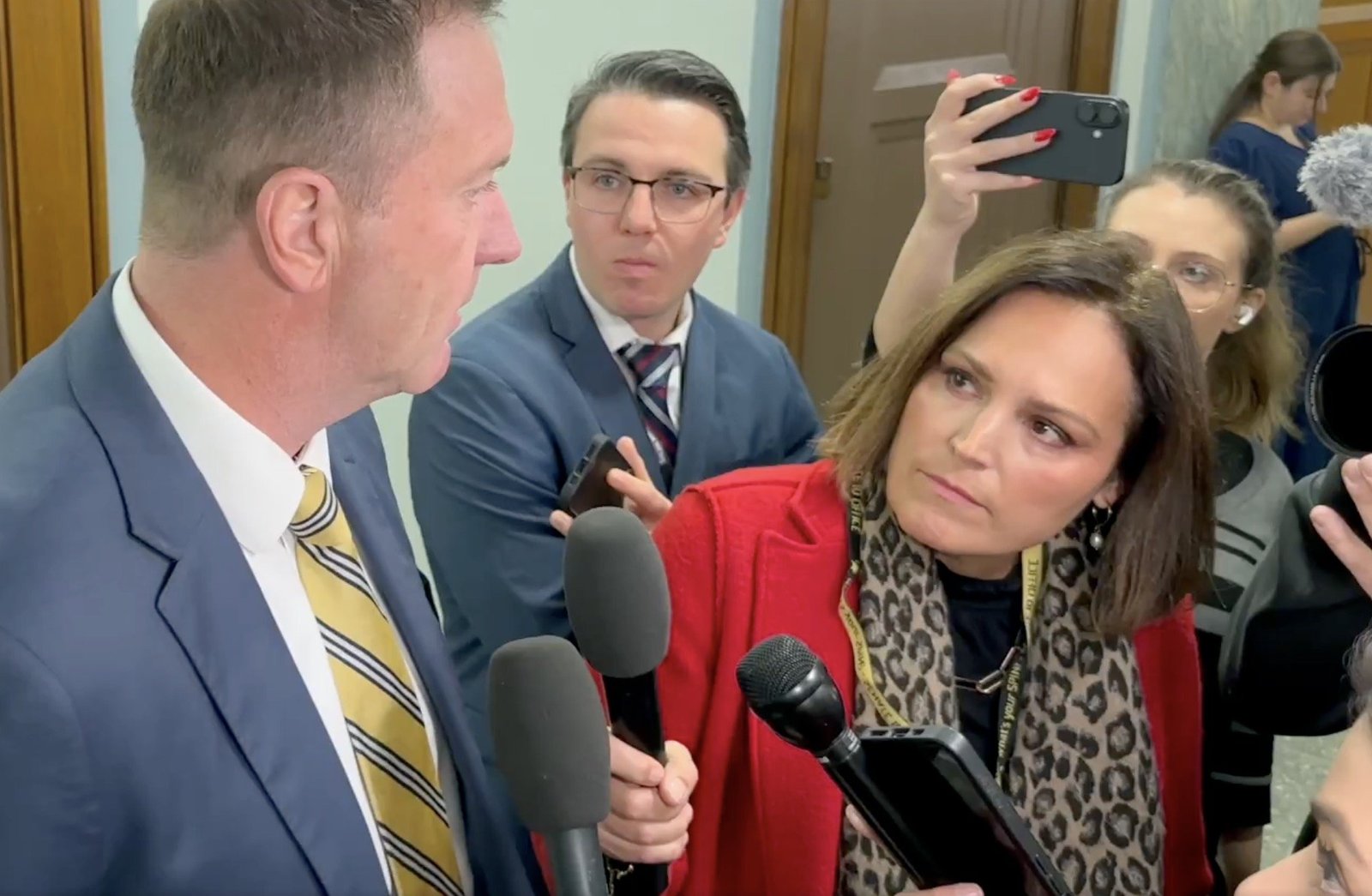 A journalist interviews a politician surrounded by cameras and reporters in a hallway, capturing a moment of media engagement and political discourse.