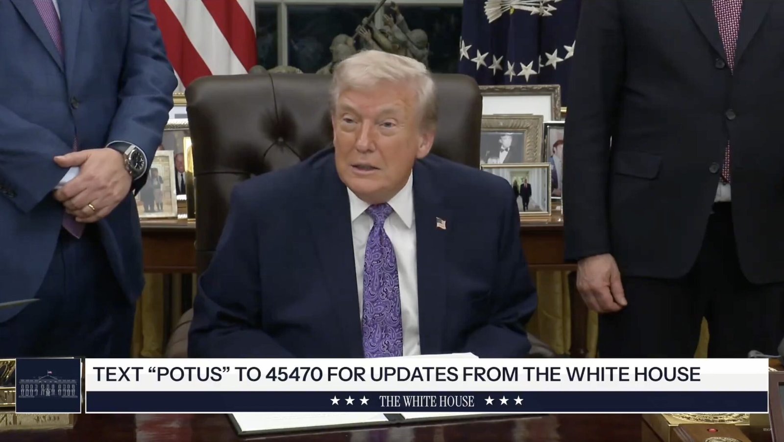 Former President Donald Trump seated at the Resolute Desk in the Oval Office, discussing updates with officials while promoting text alerts from the White House.