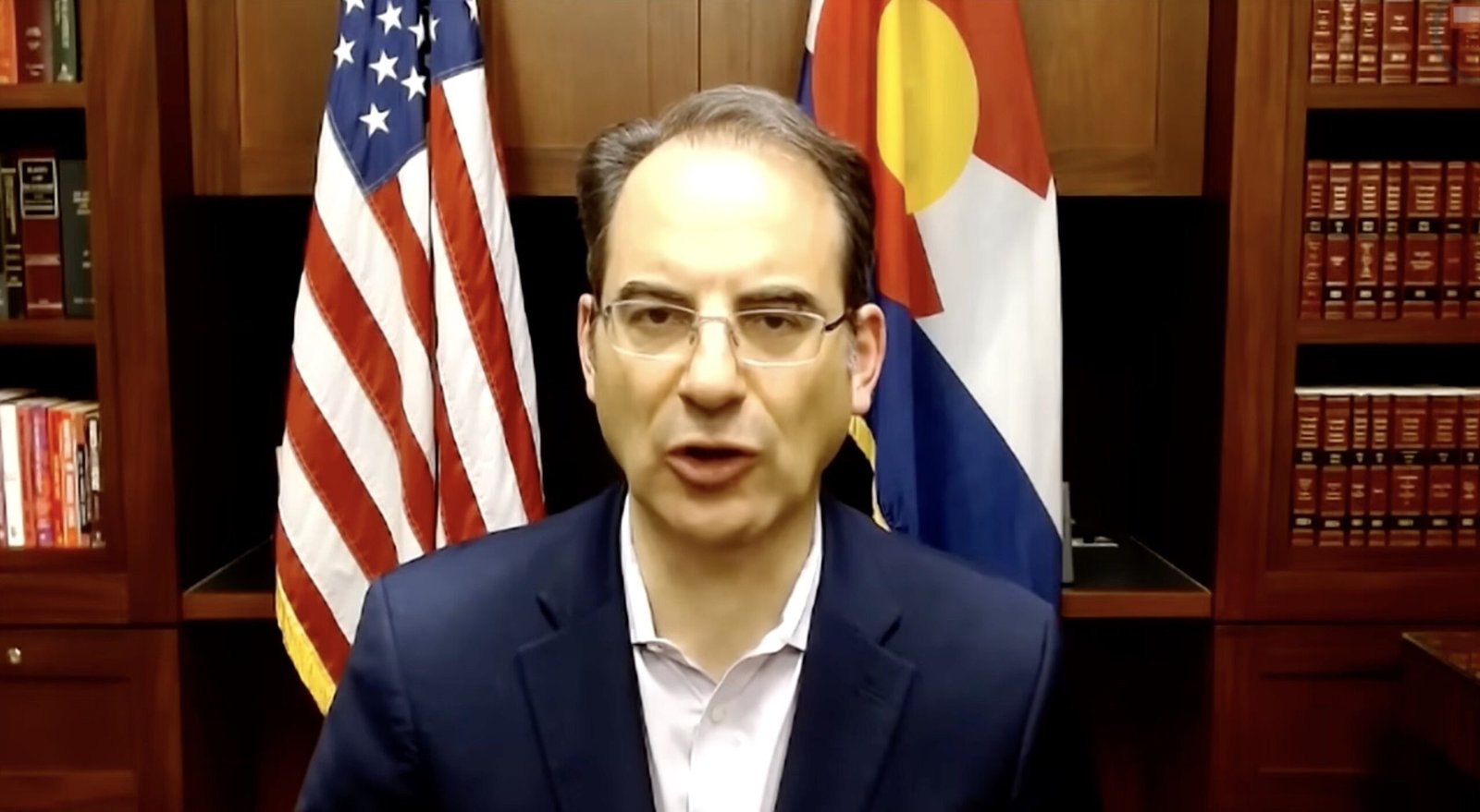 Man speaking in front of American and Colorado flags, with a bookshelf in the background, conveying a formal message or announcement.