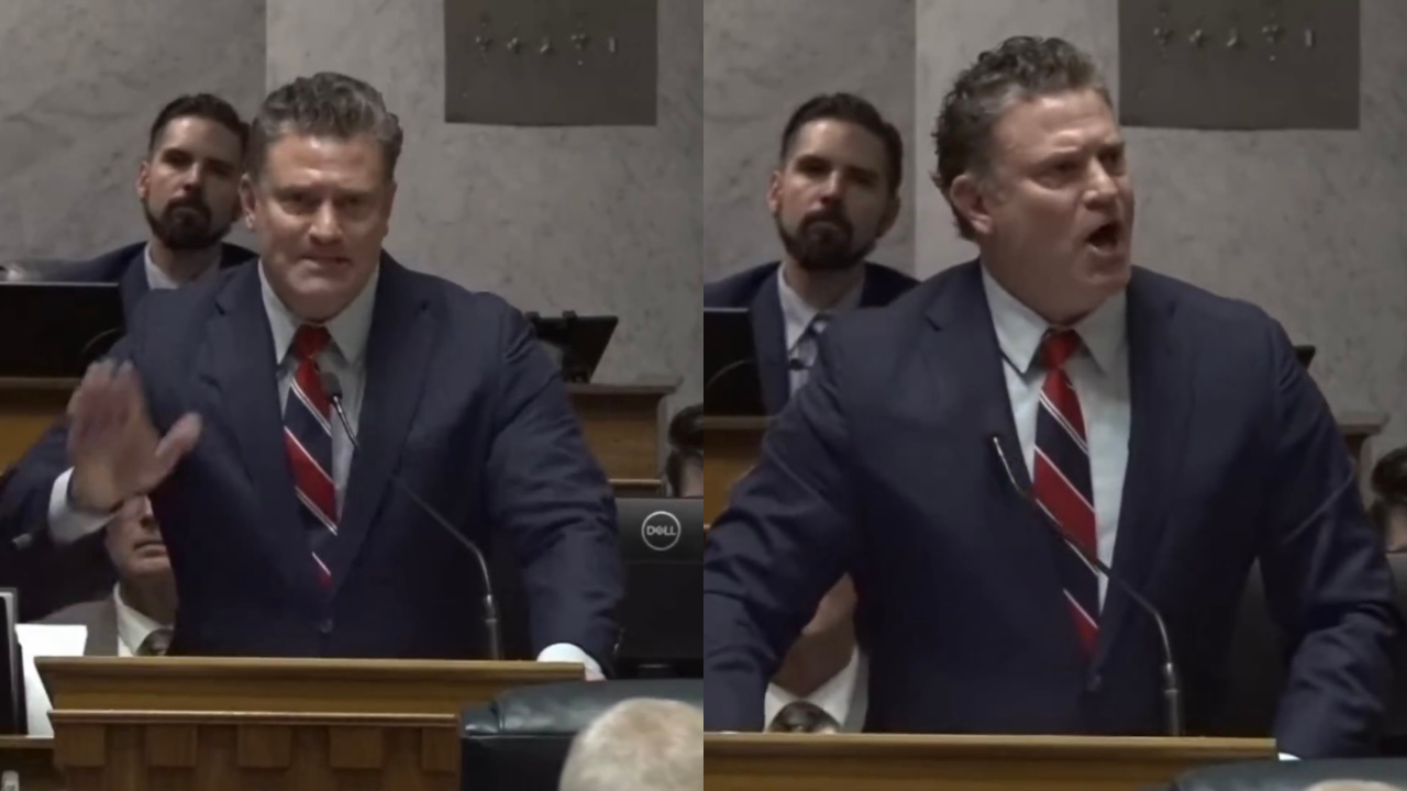 A man in a suit passionately speaks at a podium during a legislative session, with an audience visible in the background.