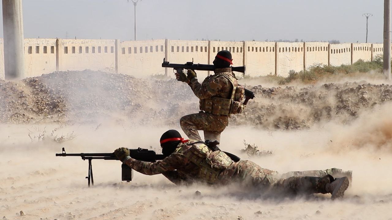 Two soldiers in tactical gear engage in training exercises, showcasing military tactics in a dusty environment with a barrier in the background.