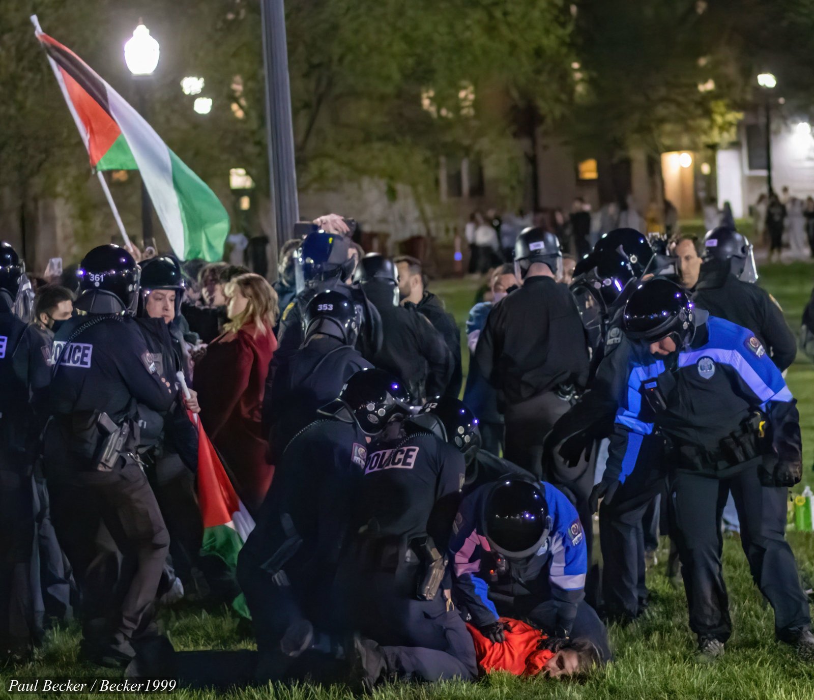 Police officers manage a crowd during a protest, with a person on the ground and a flag in the background, capturing a moment of tension and conflict.