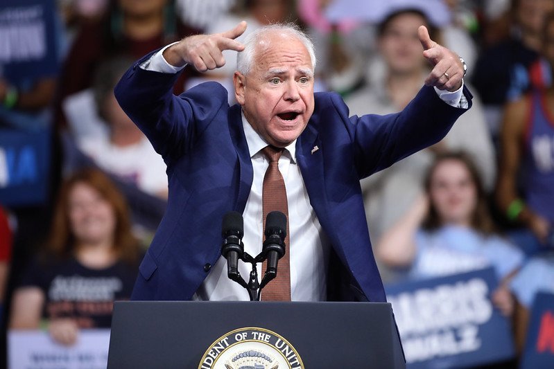 A passionate speaker gestures emphatically while addressing a crowd at a political rally, with supporters holding signs in the background.