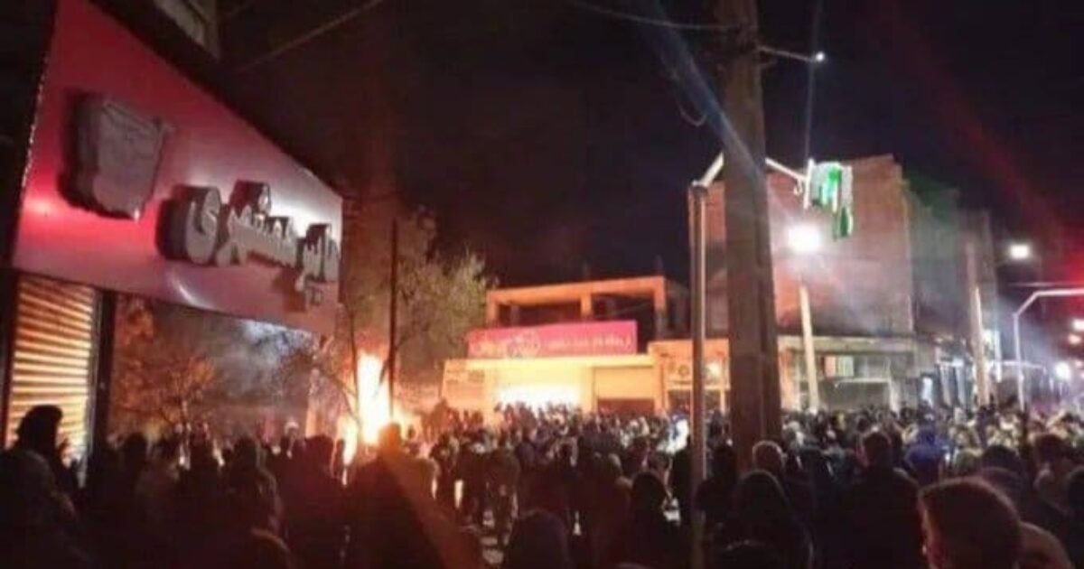 Crowd gathered in a street at night, with a fire burning and buildings illuminated by streetlights, indicating a significant public event or protest.