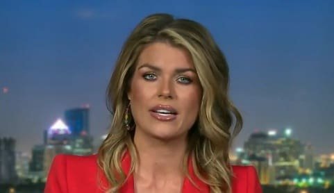 News anchor with long blonde hair wearing a red blazer, presenting against a city skyline backdrop at dusk.