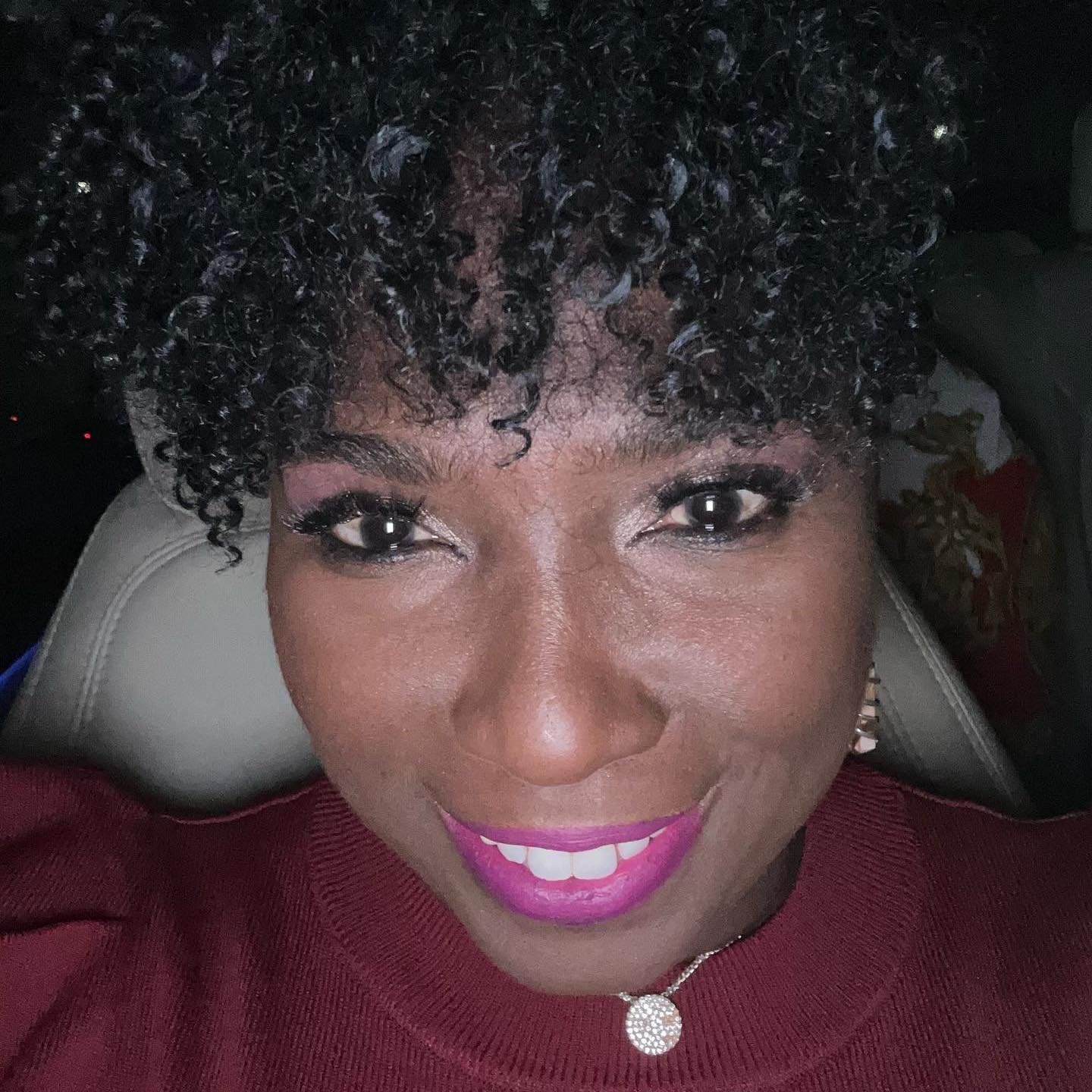 Close-up portrait of a woman with curly hair, bright pink lipstick, and a necklace, smiling in a car setting.