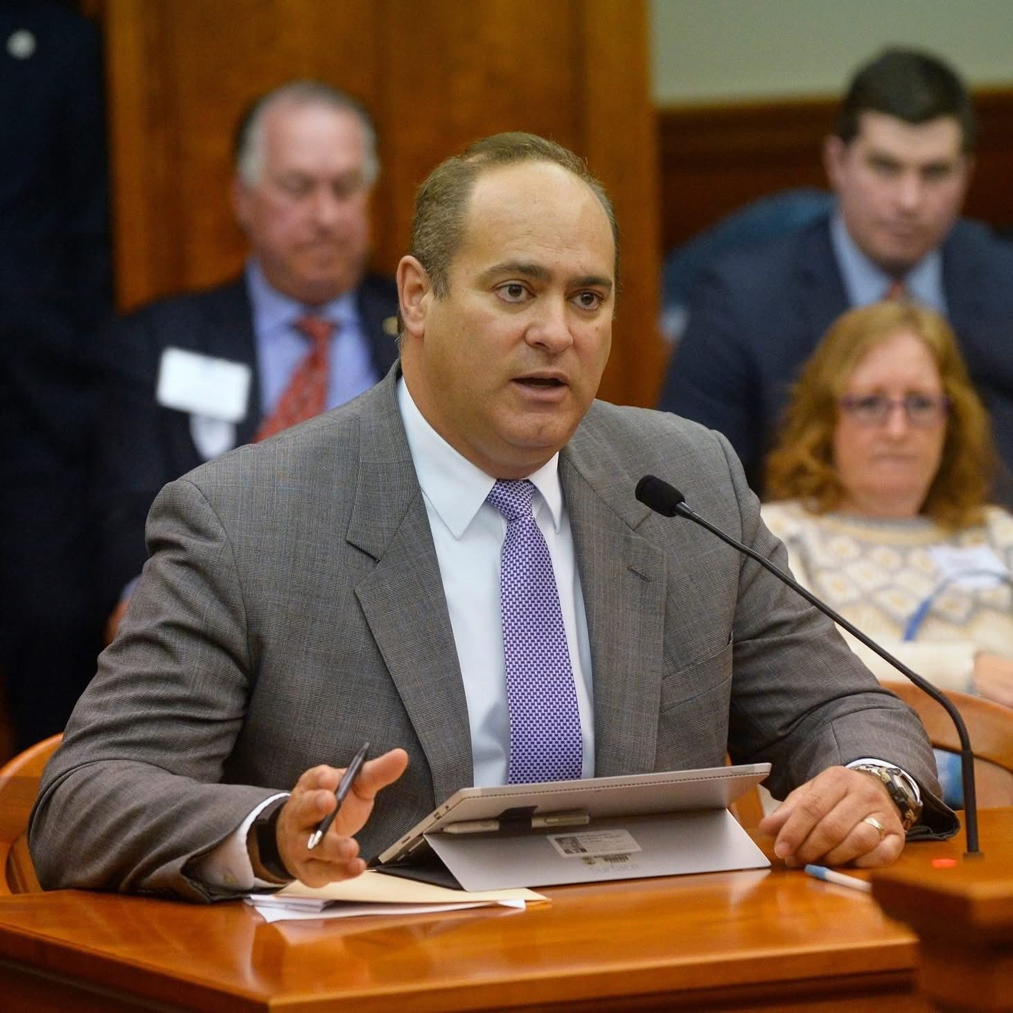 Individual in a suit speaking at a legislative hearing, with attendees seated in the background, highlighting civic engagement and public discourse.