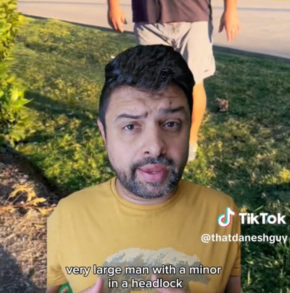 Man in a yellow shirt discusses a large individual applying a headlock on a minor, set in an outdoor environment.