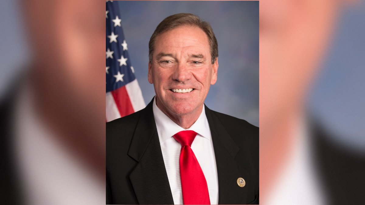 Portrait of a smiling man in a black suit and red tie, standing in front of an American flag backdrop.