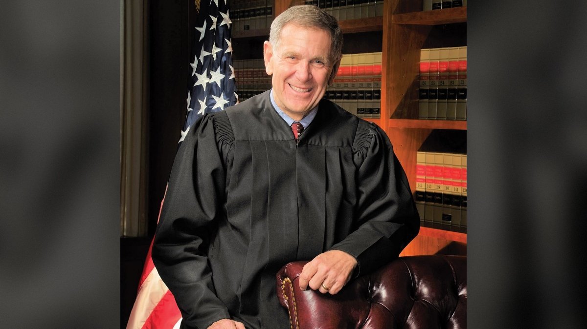 Portrait of a smiling judge in a black robe, seated in a leather chair with law books and an American flag in the background.