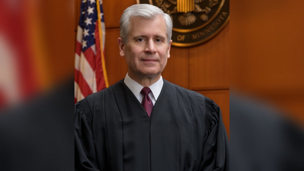 Portrait of a judge in a black robe standing in a courtroom with American flags and a seal of Minnesota in the background.