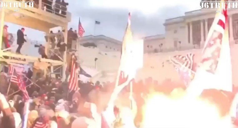 Crowd of protesters at a rally, waving flags and gathered near a building, with smoke and flames visible in the foreground.