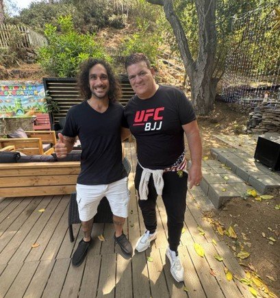 Two men pose together outdoors, one wearing a UFC BJJ shirt, in a casual setting with greenery and wooden furniture in the background.