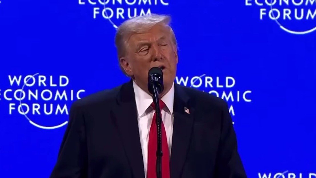 Donald Trump speaking at the World Economic Forum, addressing global economic issues with a microphone in front of a blue backdrop featuring the event's logo.