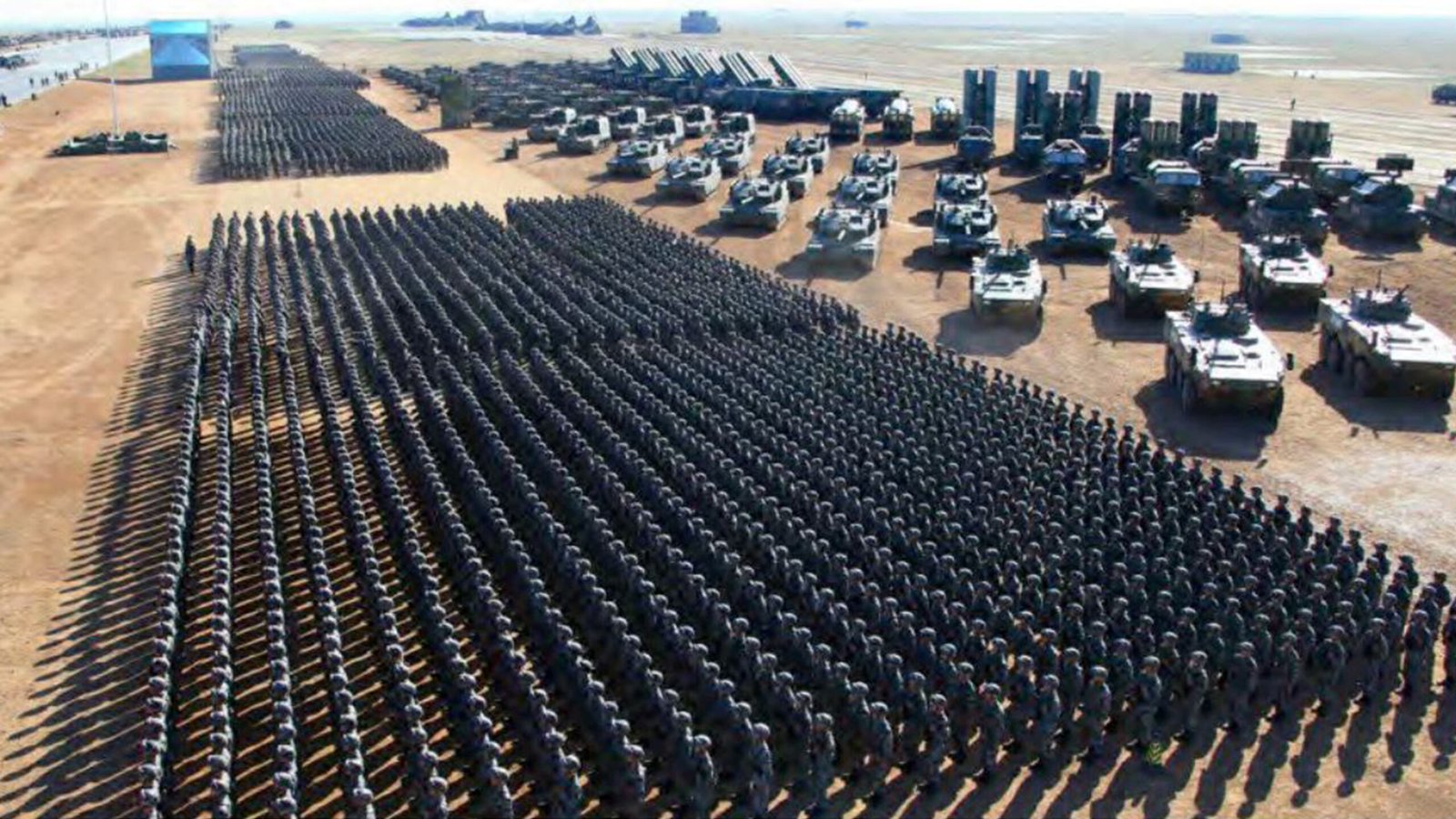 Aerial view of a large military formation with soldiers and armored vehicles arranged in rows on a desert landscape.