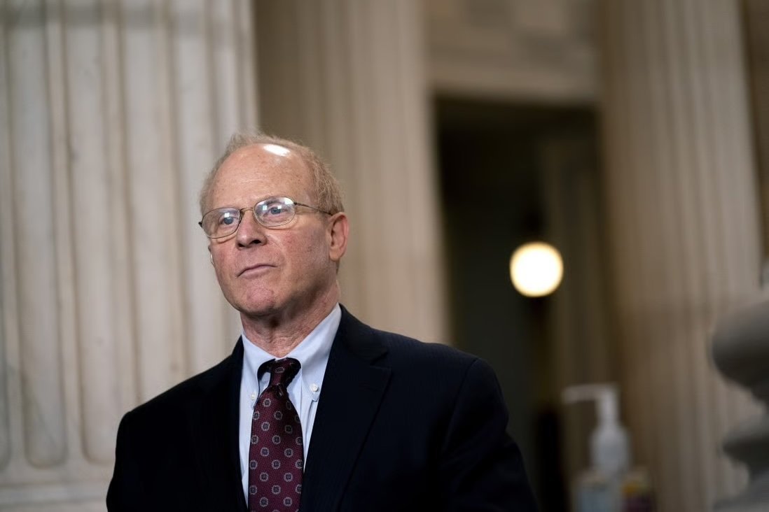 Man in a suit and glasses standing in a formal setting with pillars in the background, looking thoughtfully ahead.