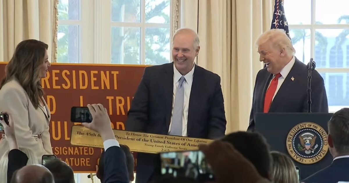 Donald Trump participates in a ceremonial event, smiling while presenting a commemorative plaque to a guest, surrounded by attendees capturing the moment on their phones.