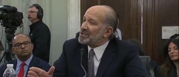 A man in a suit testifies during a Senate hearing, with cameras and an audience visible in the background.