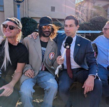 Group of four men posing for a photo outdoors, two wearing sunglasses, one in a blazer, and another with long hair, while one holds a microphone.