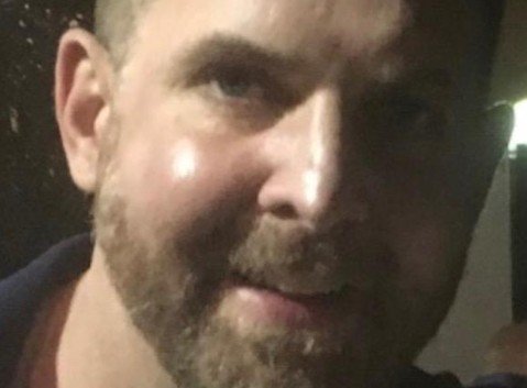 Close-up of a smiling man with a beard, captured in low light, showcasing facial features and a friendly expression.