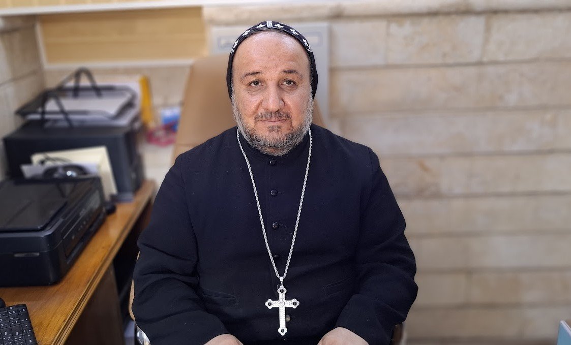 Portrait of a religious leader in traditional attire, seated at a desk with office supplies and a printer, exuding a calm and authoritative presence.