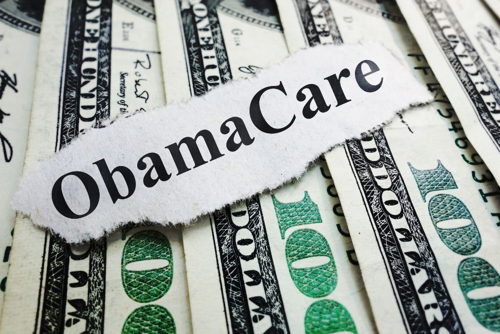 A torn piece of paper with the word ObamaCare on a background of hundred-dollar bills, symbolizing healthcare costs and financial aspects of the Affordable Care Act.