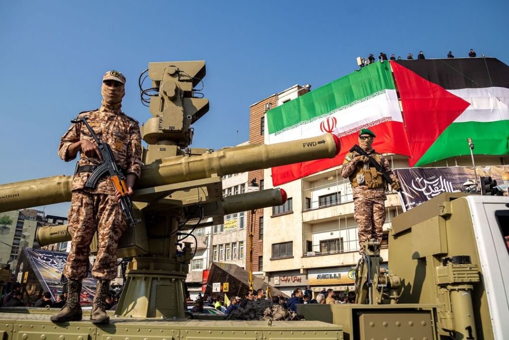 Military personnel stand on armored vehicles beneath a large flag representing Iran and Palestine during a public demonstration, showcasing military presence and solidarity.