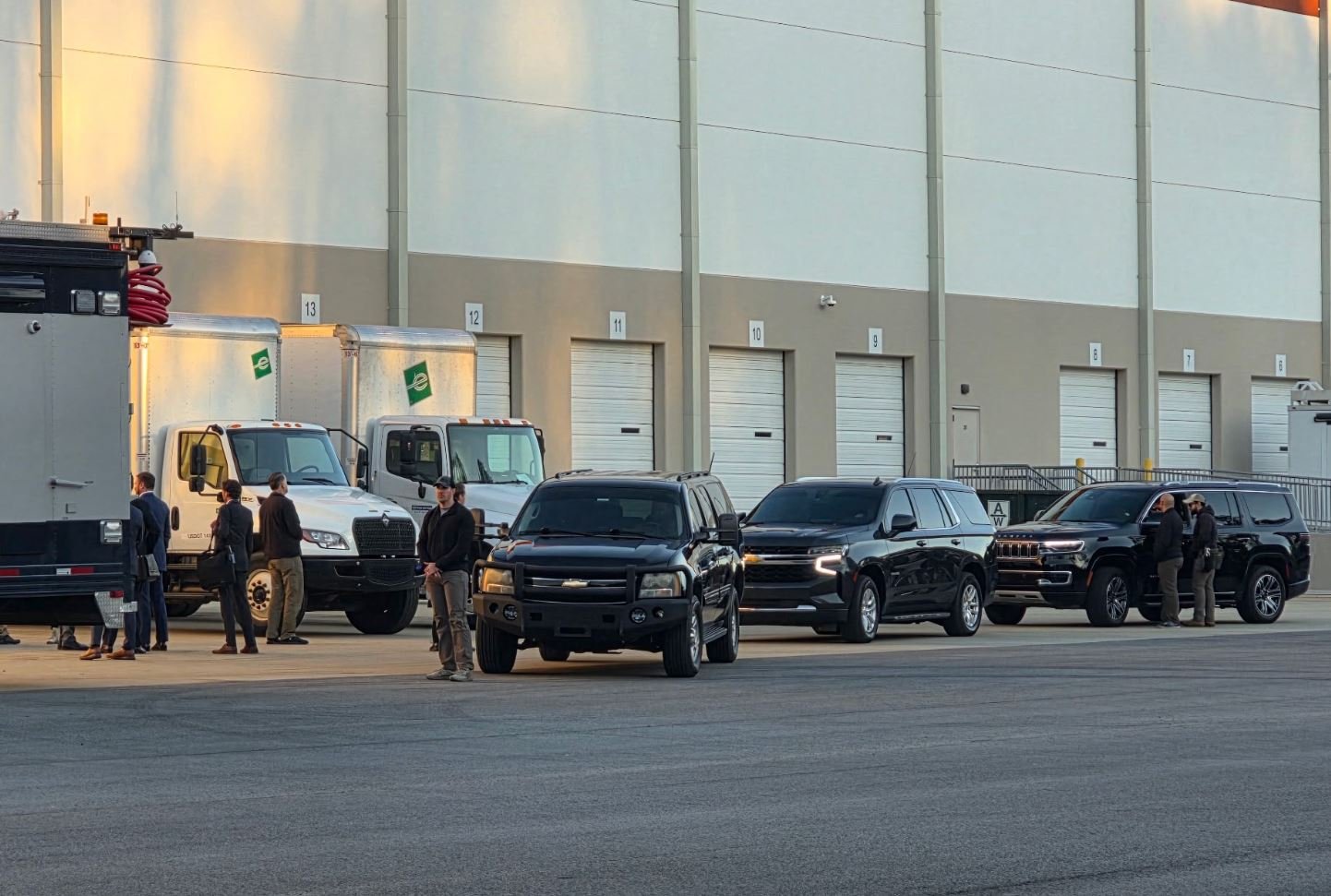 Several SUVs and trucks parked outside a warehouse, with people standing nearby, during early morning light.