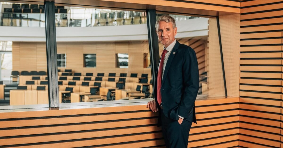 A professional man in a suit stands by a window, overlooking a modern conference room with wooden accents and empty seating.