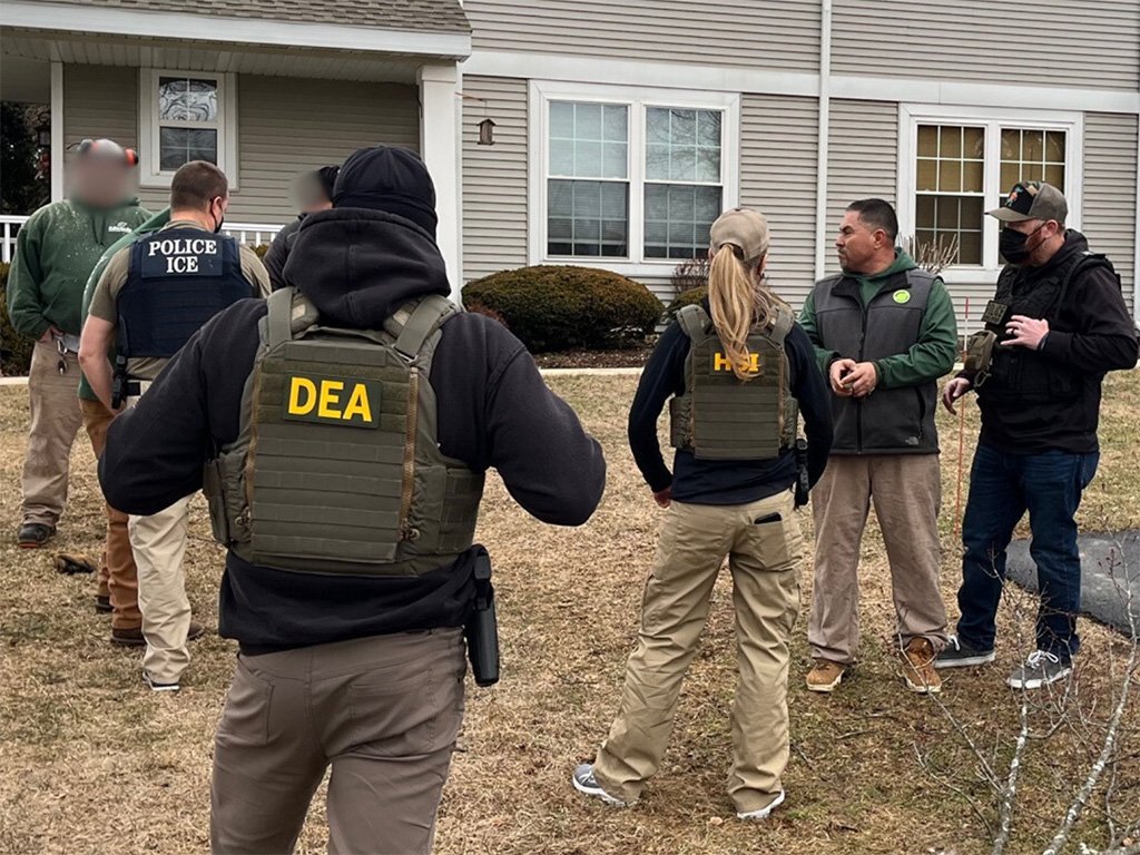 Law enforcement agents from DEA and ICE conduct an operation outside a residential home, discussing details in tactical gear.