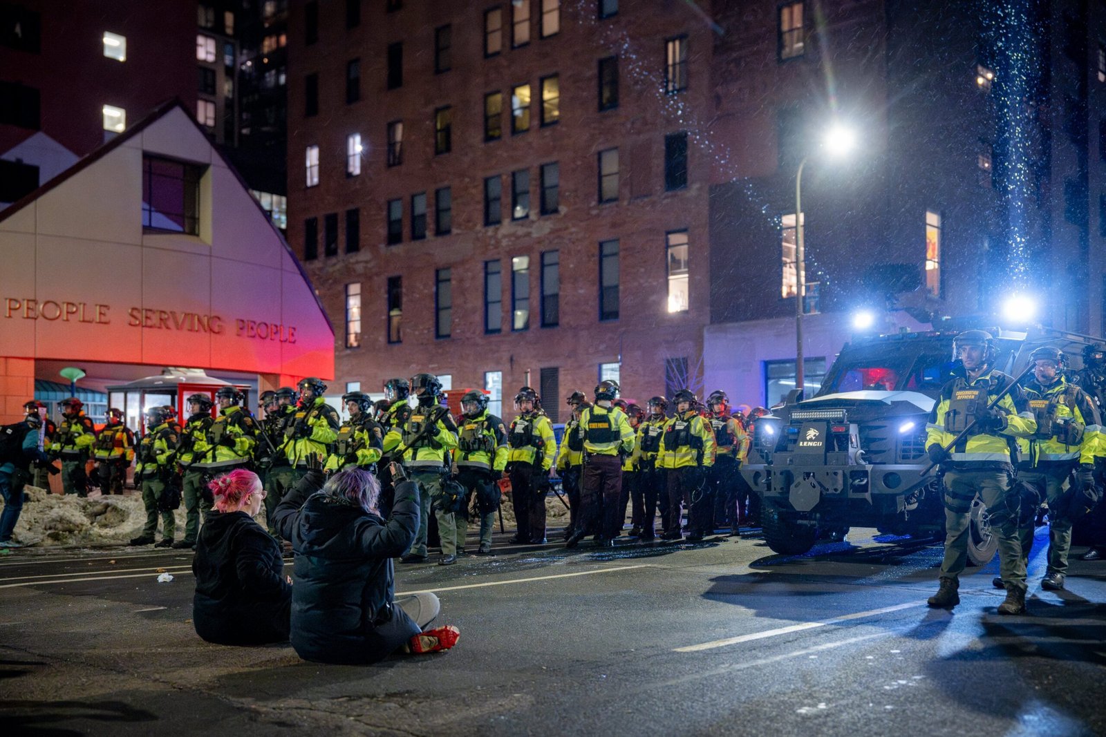 Police in riot gear face protesters sitting on the street outside a building labeled "People Serving People" during a nighttime demonstration.