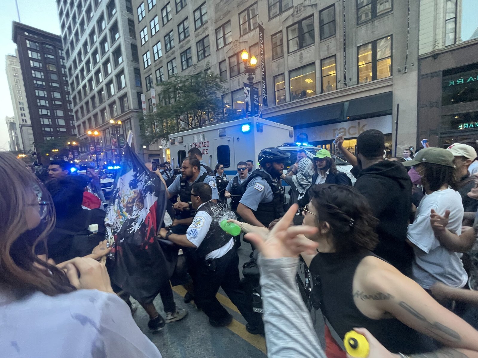 Crowd of protesters gather in downtown Chicago, interacting with police officers amid flashing lights and a visible emergency vehicle.