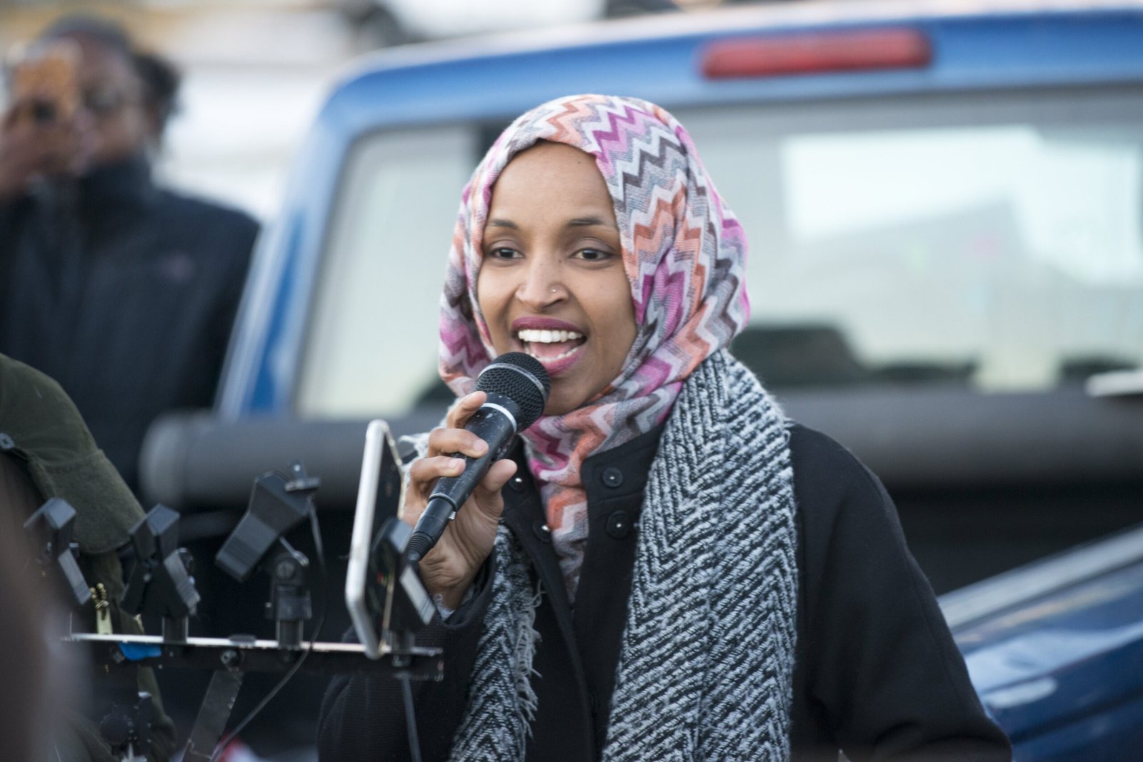 Woman in a colorful hijab speaks passionately into a microphone at a public event, surrounded by an engaged audience and a blue pickup truck in the background.