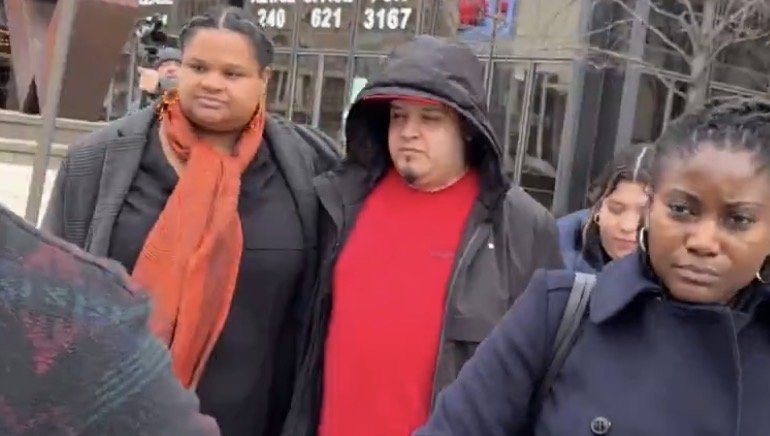 A group of individuals stands outside a building, with two people prominently featured in the foreground, one wearing an orange scarf and another in a red shirt.