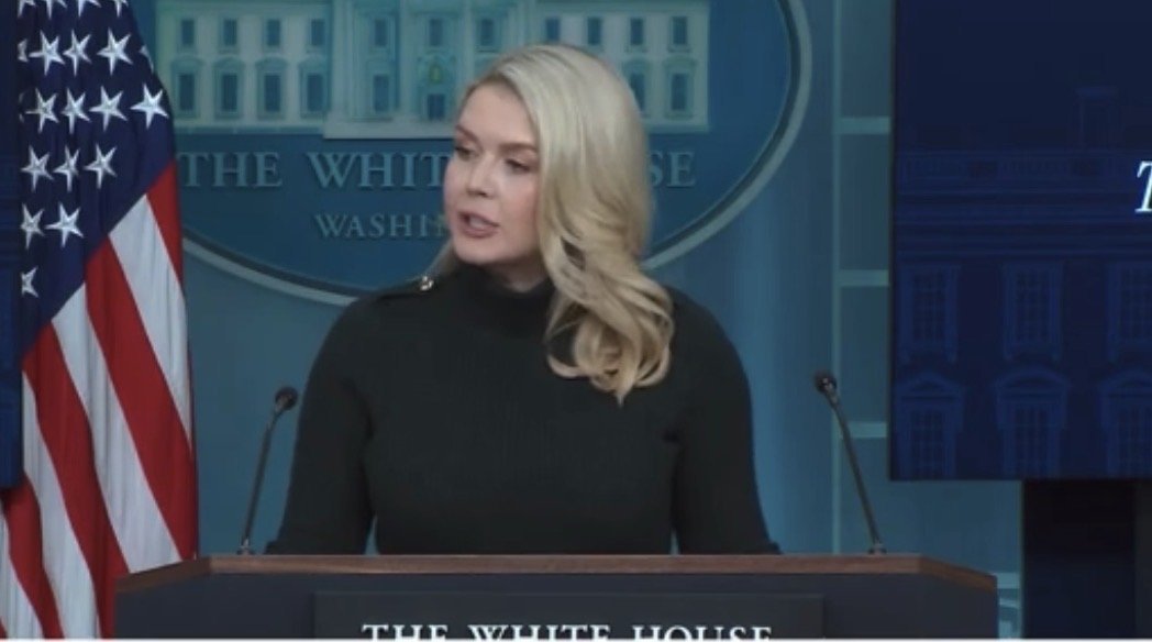 Press secretary delivers a statement at the White House briefing room, with the American flag in the background and the White House seal visible.