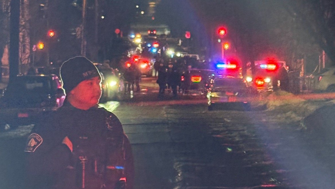 Police officer on duty in a dimly lit street with emergency vehicles and flashing lights in the background, indicating a potential incident or investigation.
