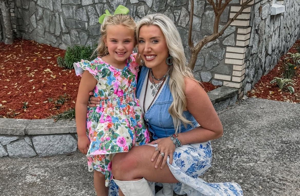 A smiling woman in a denim top and floral skirt poses with a young girl in a colorful dress outside a stone wall.