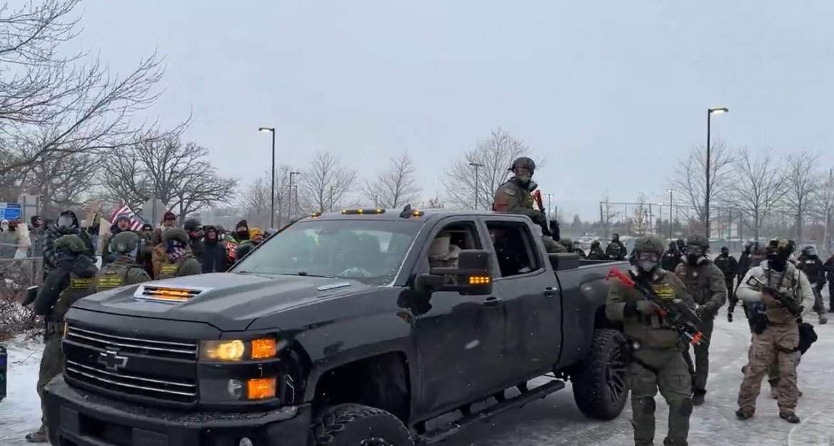 Armed individuals in tactical gear gather around a black pickup truck during a winter protest, with flags and signs visible in the background.