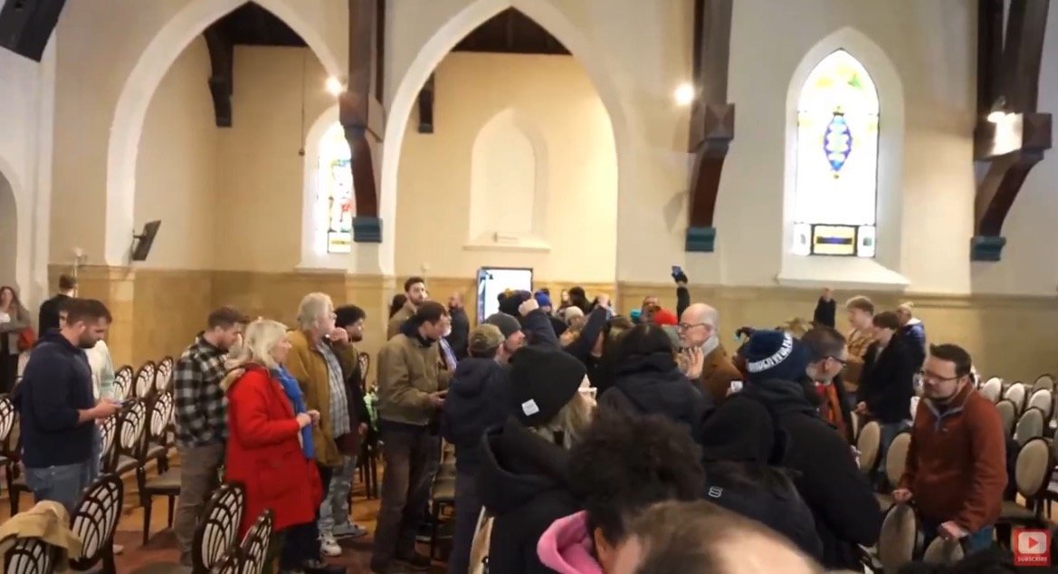A crowd of people gathers inside a church with stained glass windows, engaging in conversation and using smartphones.