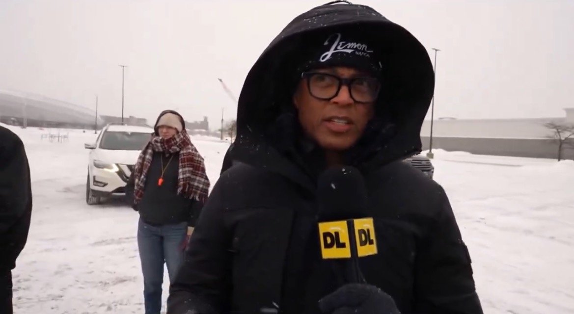News reporter in winter attire stands outside in snowy conditions, providing live coverage while a woman and a vehicle are visible in the background.