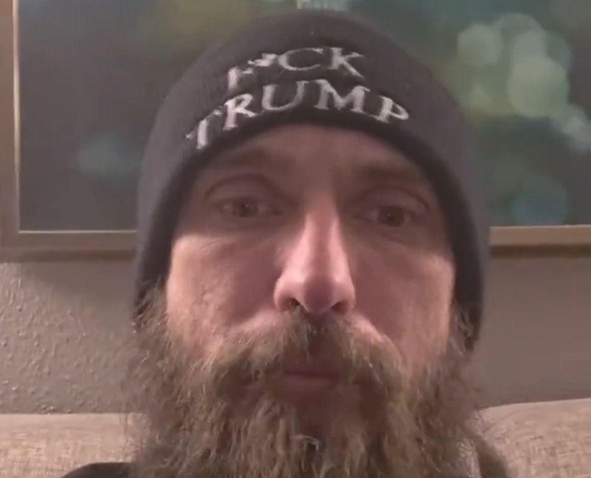 Man with a beard wearing a black beanie with anti-Trump message, sitting indoors in a softly lit room.
