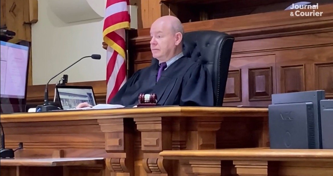 Judge presiding over a courtroom session, with an American flag in the background and a gavel on the desk, focused on a computer screen.