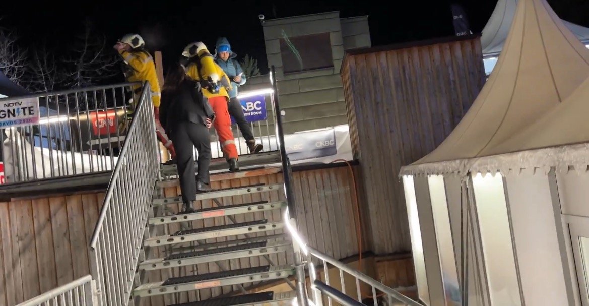 Skiers and a woman in formal attire ascend a lit staircase at a winter sports venue during nighttime, showcasing a blend of leisure and professionalism.